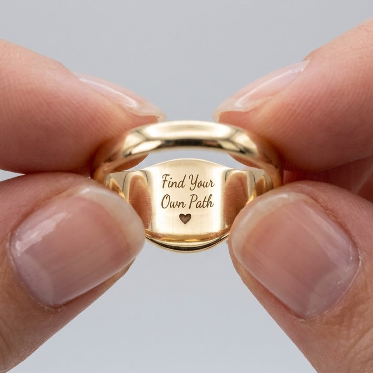 A gold signet ring held between two fingers, featuring the engraved text 'Find Your Own Path' and a small heart on the inner band, set against a neutral gray background.