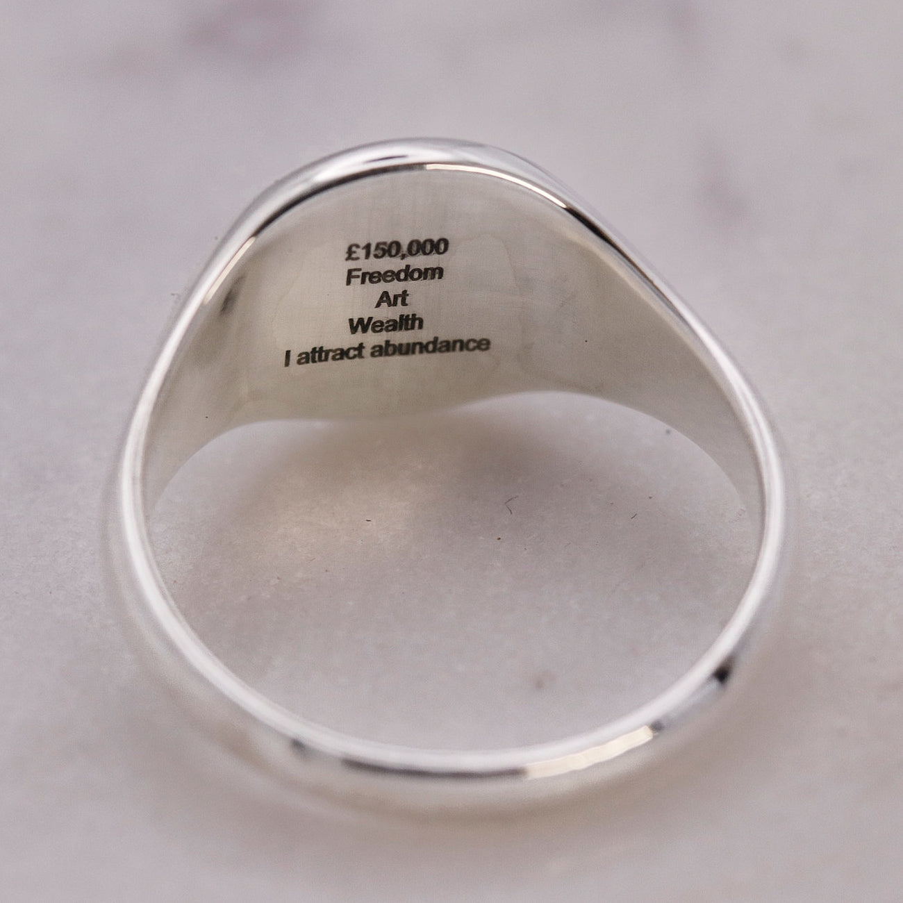 A silver signet ring with engraved black text inside the band reading '£150,000 Freedom Art Wealth I attract abundance', shown on a light marble surface.