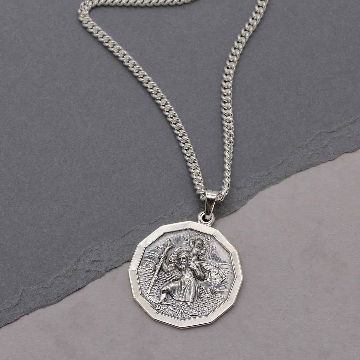 A silver chain necklace with an octagonal pendant featuring an embossed religious figure, displayed on a split gray background.