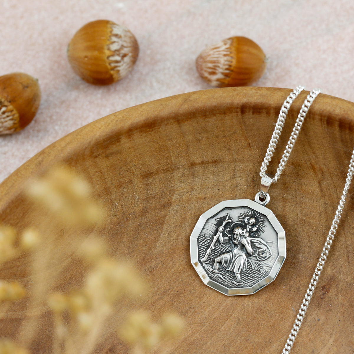 A silver pendant necklace with an octagonal medallion featuring an engraved figure, displayed on a wooden plate with acorns in the background.