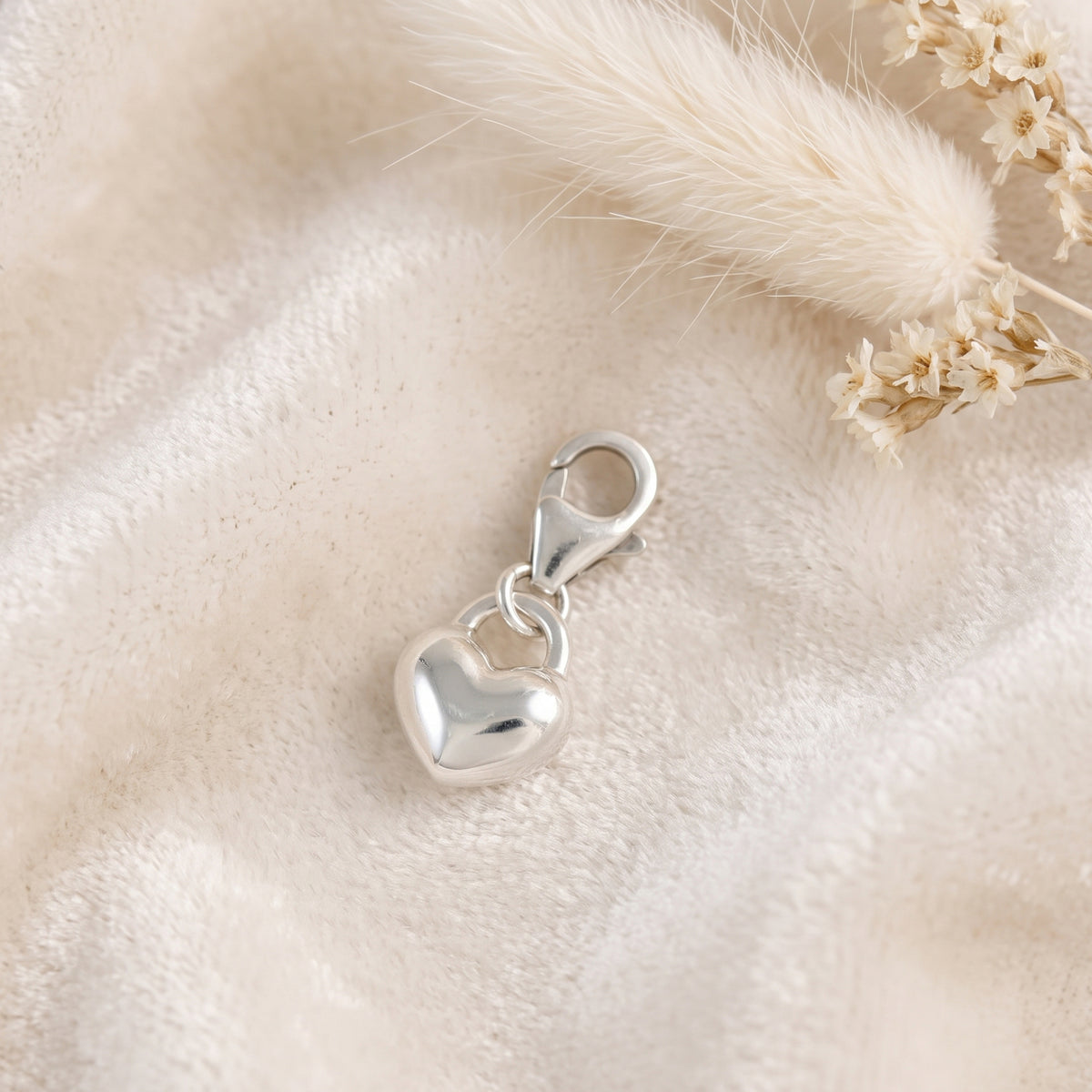 A polished silver heart charm with a lobster clasp, resting on a soft beige textured fabric with dried decorative grass and small flowers.