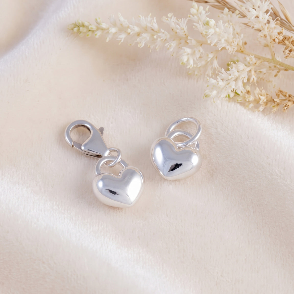 Two polished silver heart-shaped charm clasps resting on a soft cream satin fabric with dried white flowers in the corner.
