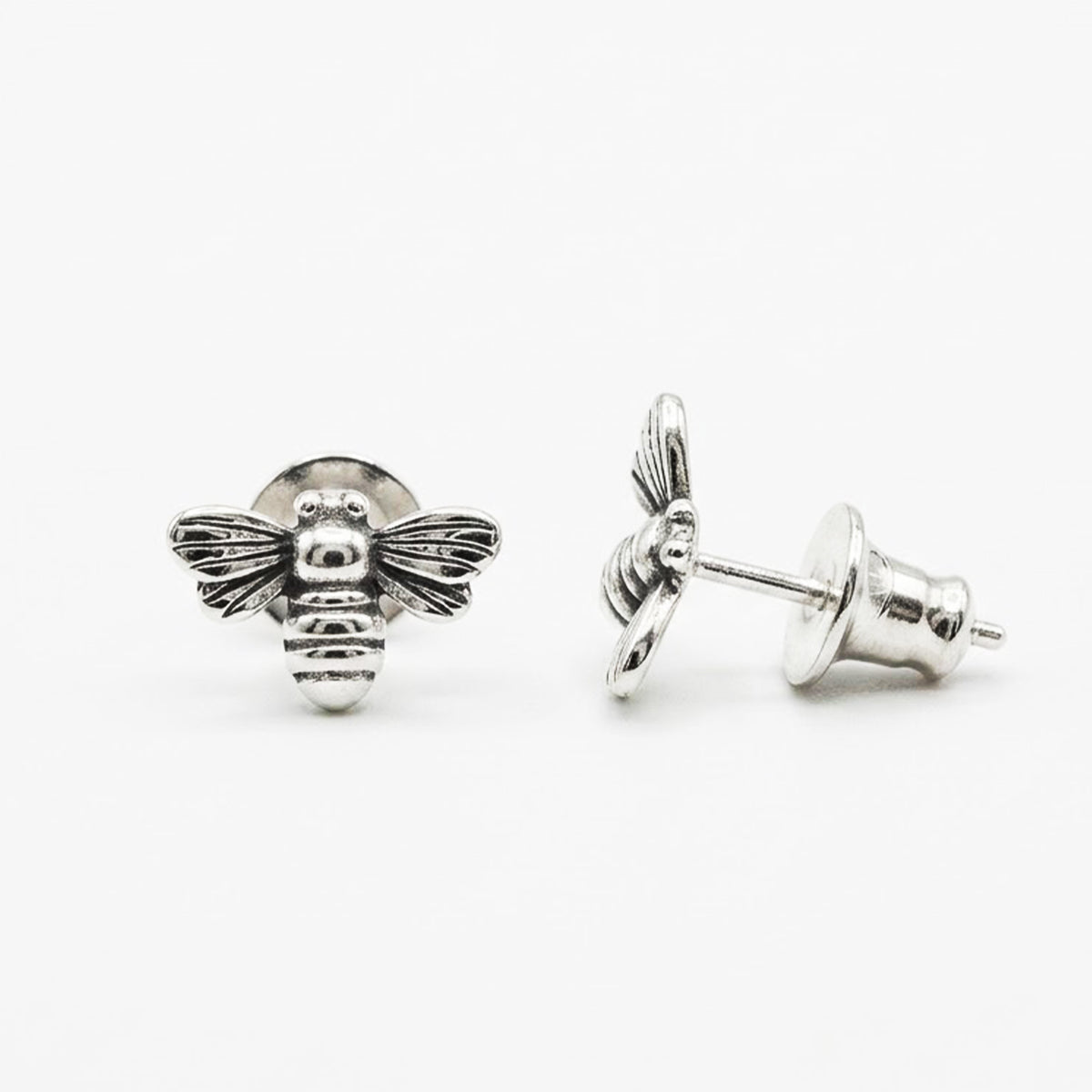 A pair of silver bee stud earrings, with one shown from the front displaying detailed wing and body carvings, and the other from the side showing the post and butterfly backing. Photographed on a clean, light gray background.