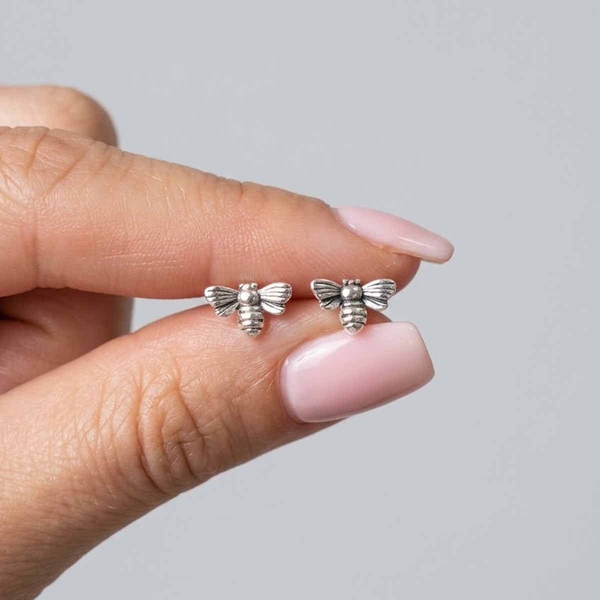 A close-up of a hand with light pink manicured nails holding a pair of small silver bee stud earrings against a plain light gray background.