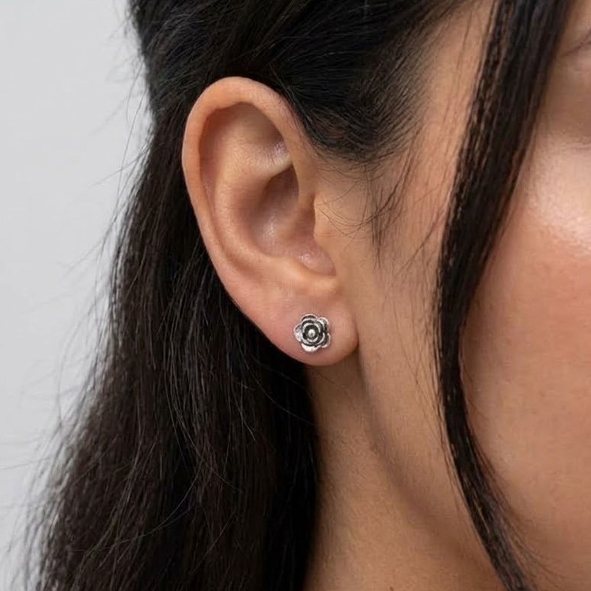 A close-up of a person's ear wearing a small silver rose-shaped stud earring against a light gray background.