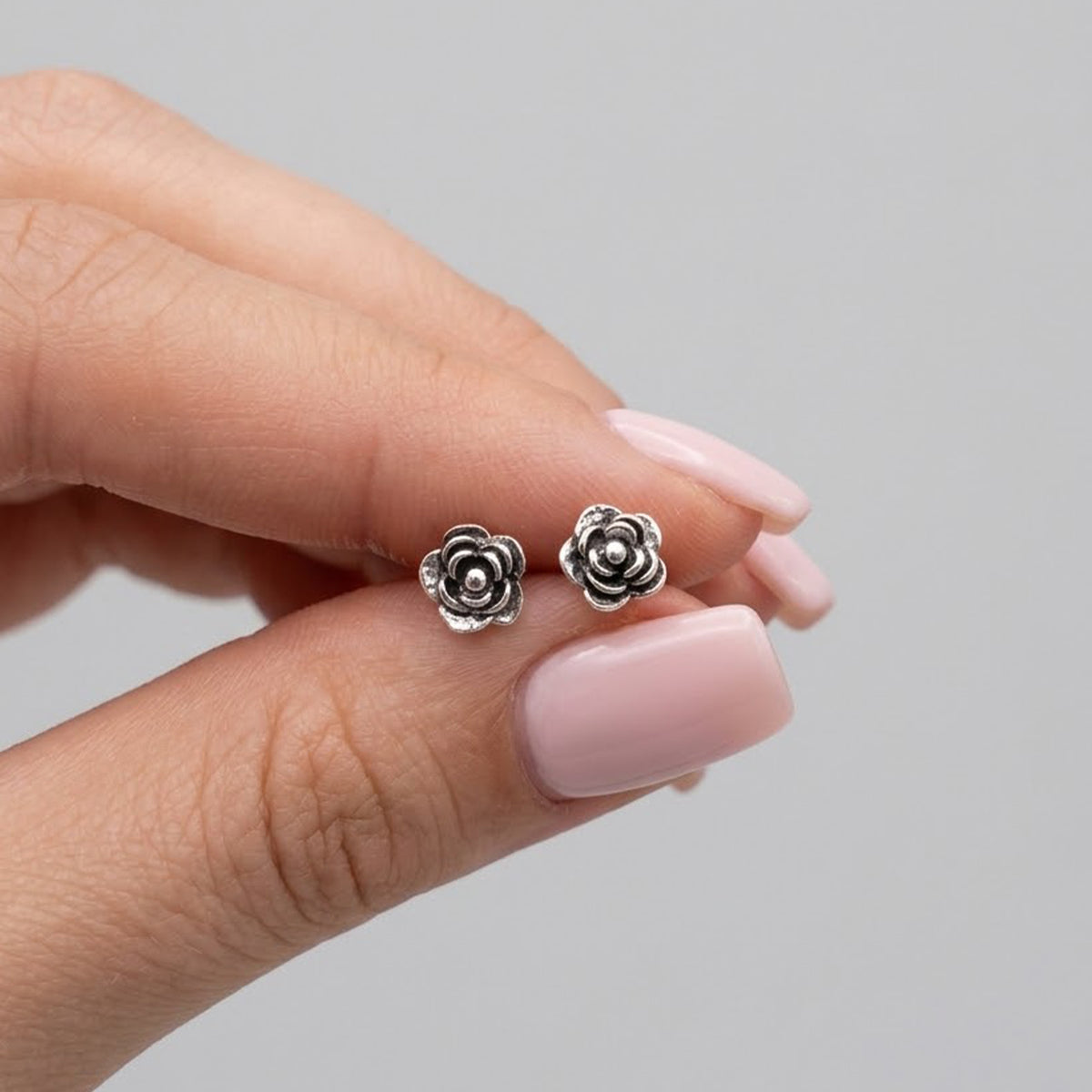 A close-up of a hand with light pink manicured nails holding a pair of small silver rose stud earrings against a plain light gray background.