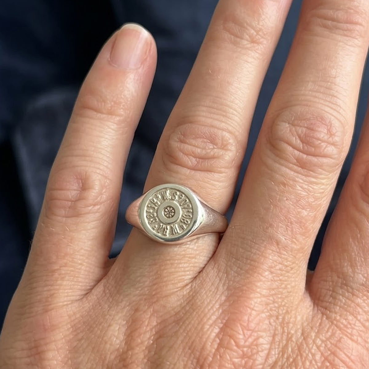 A silver signet ring with a circular, medallion-like face featuring raised lettering, shown worn on a person's ring finger against a dark navy background.