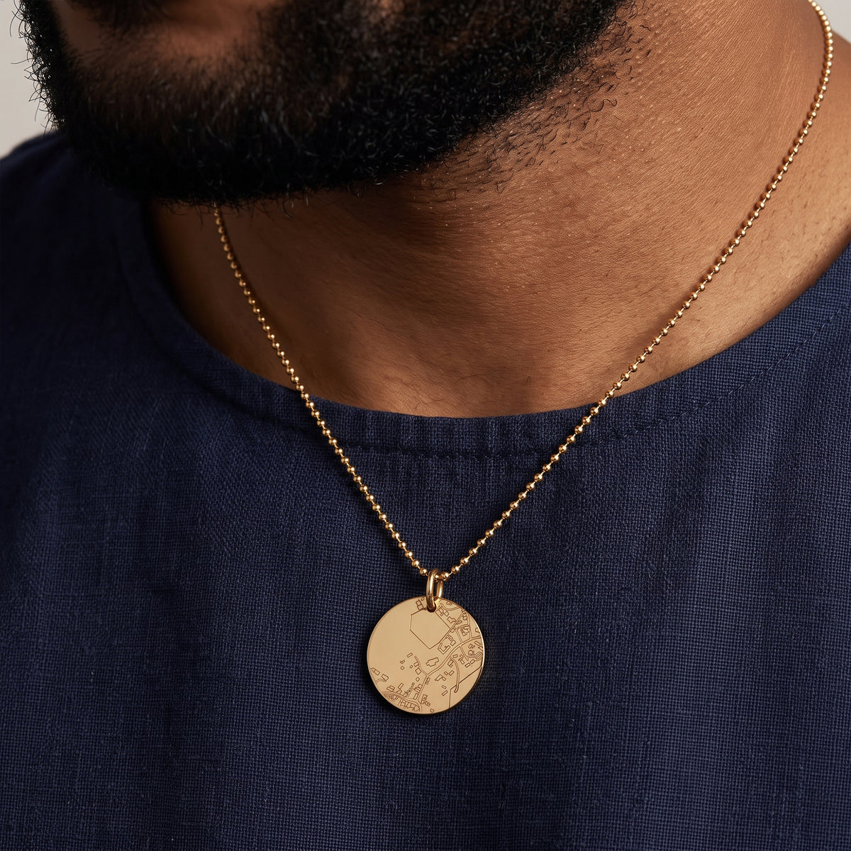 A close-up of a man wearing a gold chain necklace with a round engraved gold pendant, paired with a navy blue shirt.