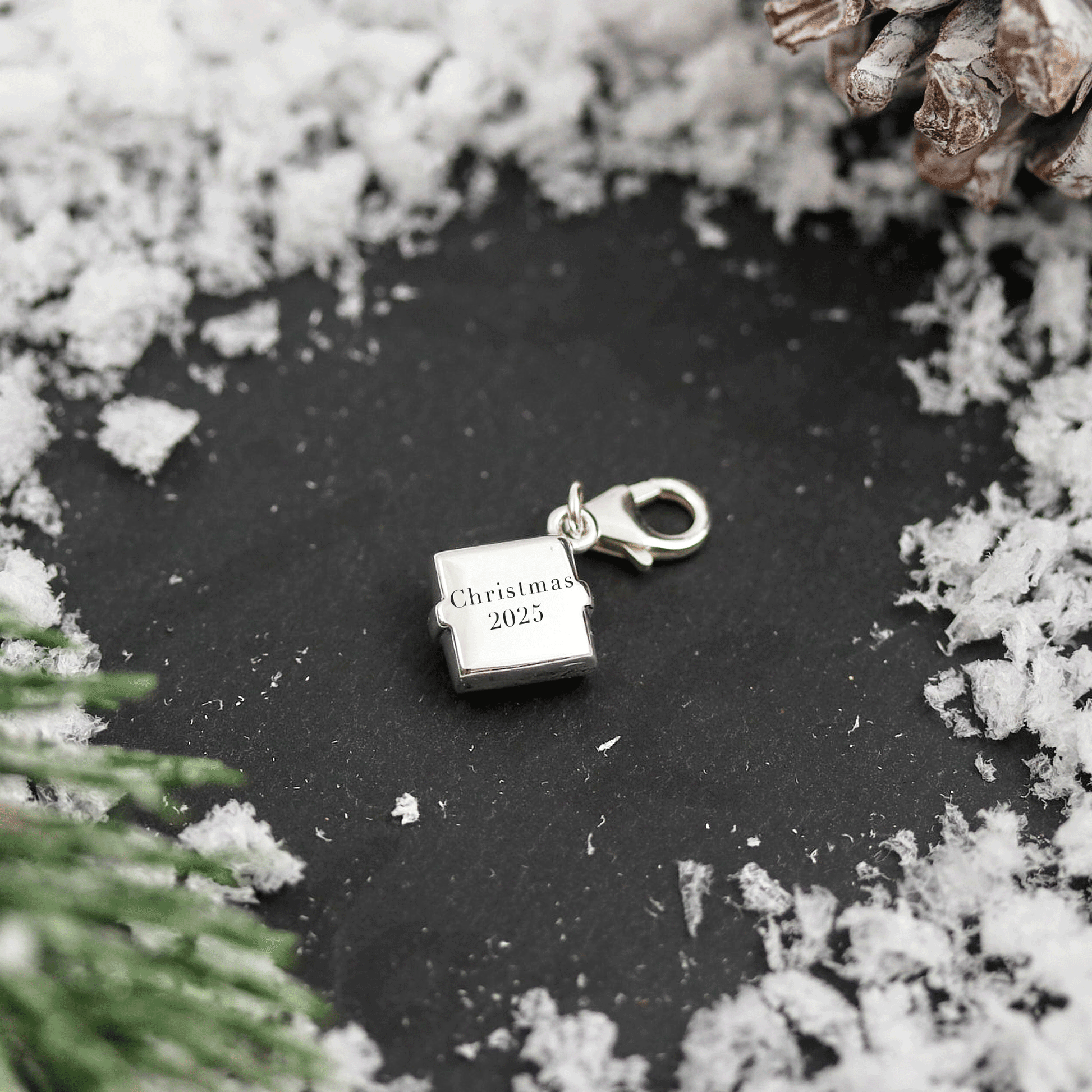 Silver present box charm with 'Christmas 2025' text on a dark surface with snow and pine branches.