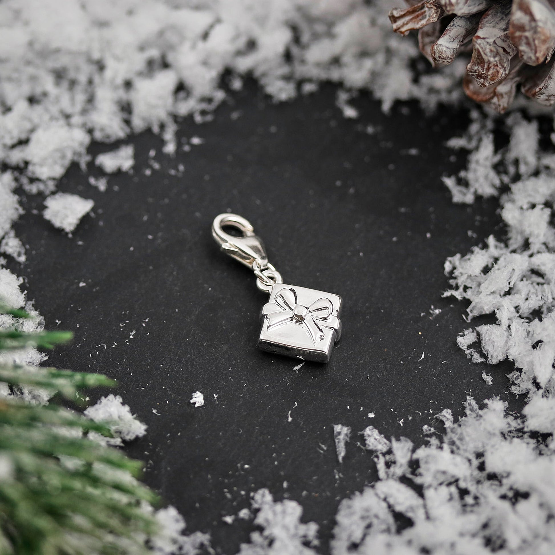 Silver charm shaped like a gift box with a bow on a dark surface surrounded by snow and pine branches.