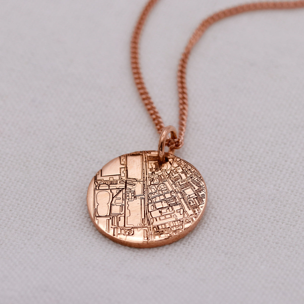 A rose gold pendant necklace featuring an engraved circular map design, displayed on a textured light gray background.