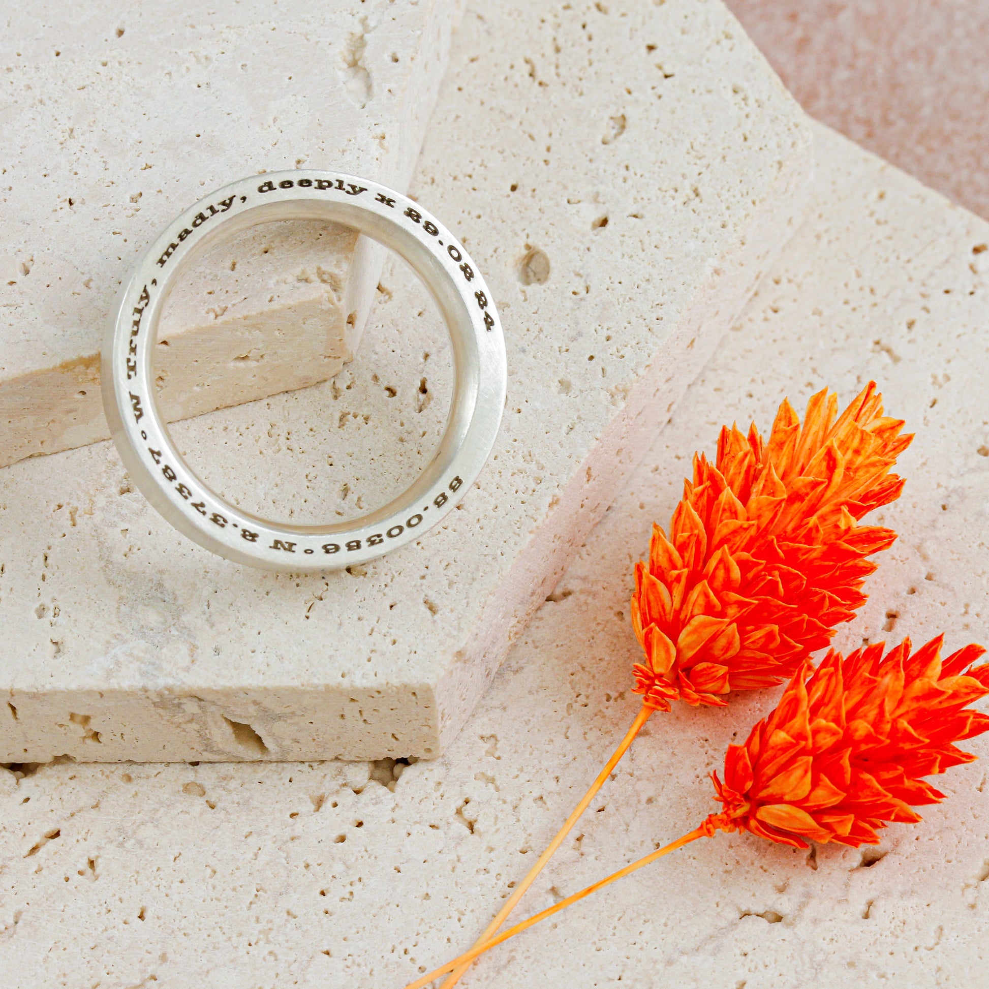Silver ring with engraved text on a textured surface with orange flowers.