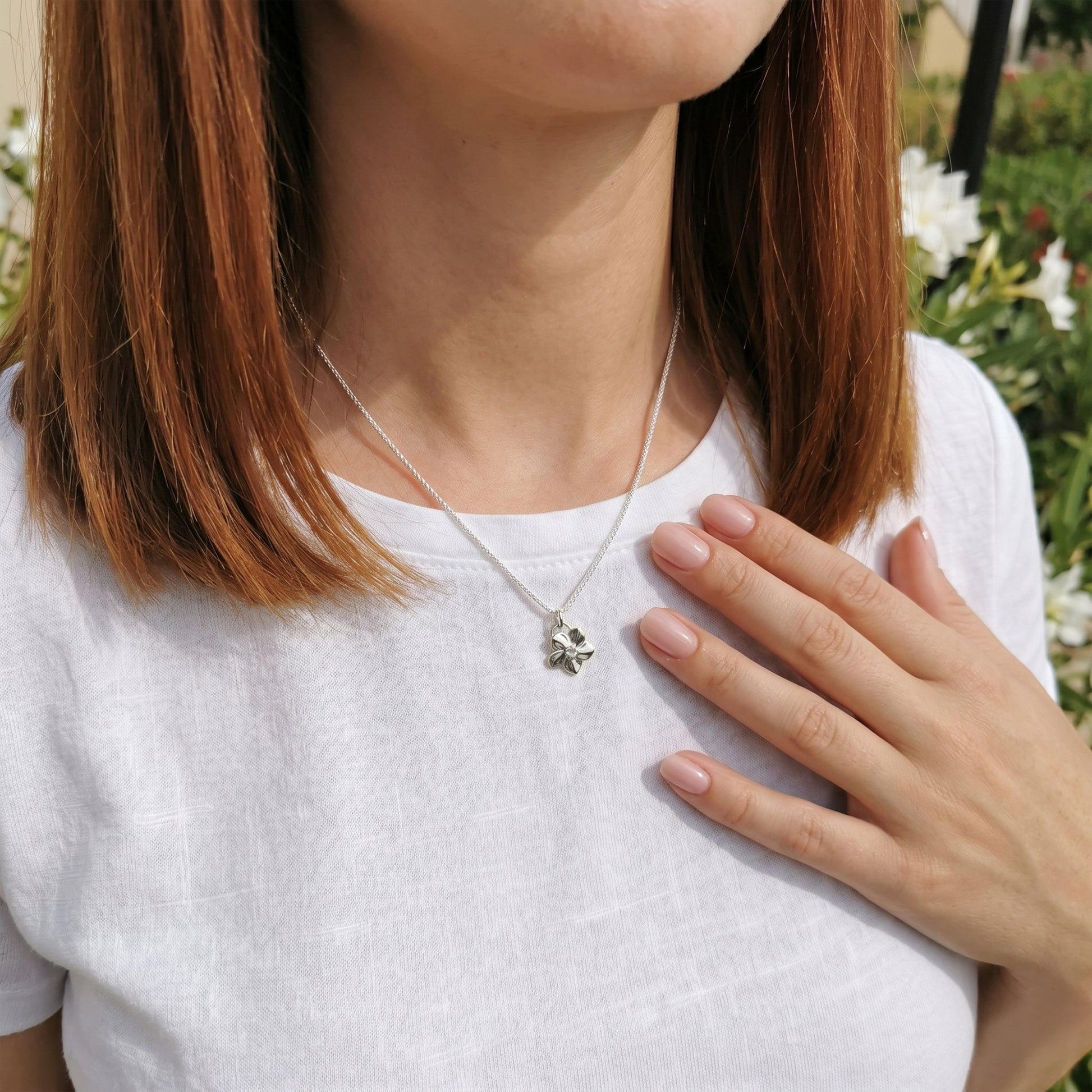 Person wearing a silver necklace with a flower pendant, standing outdoors with greenery in the background.