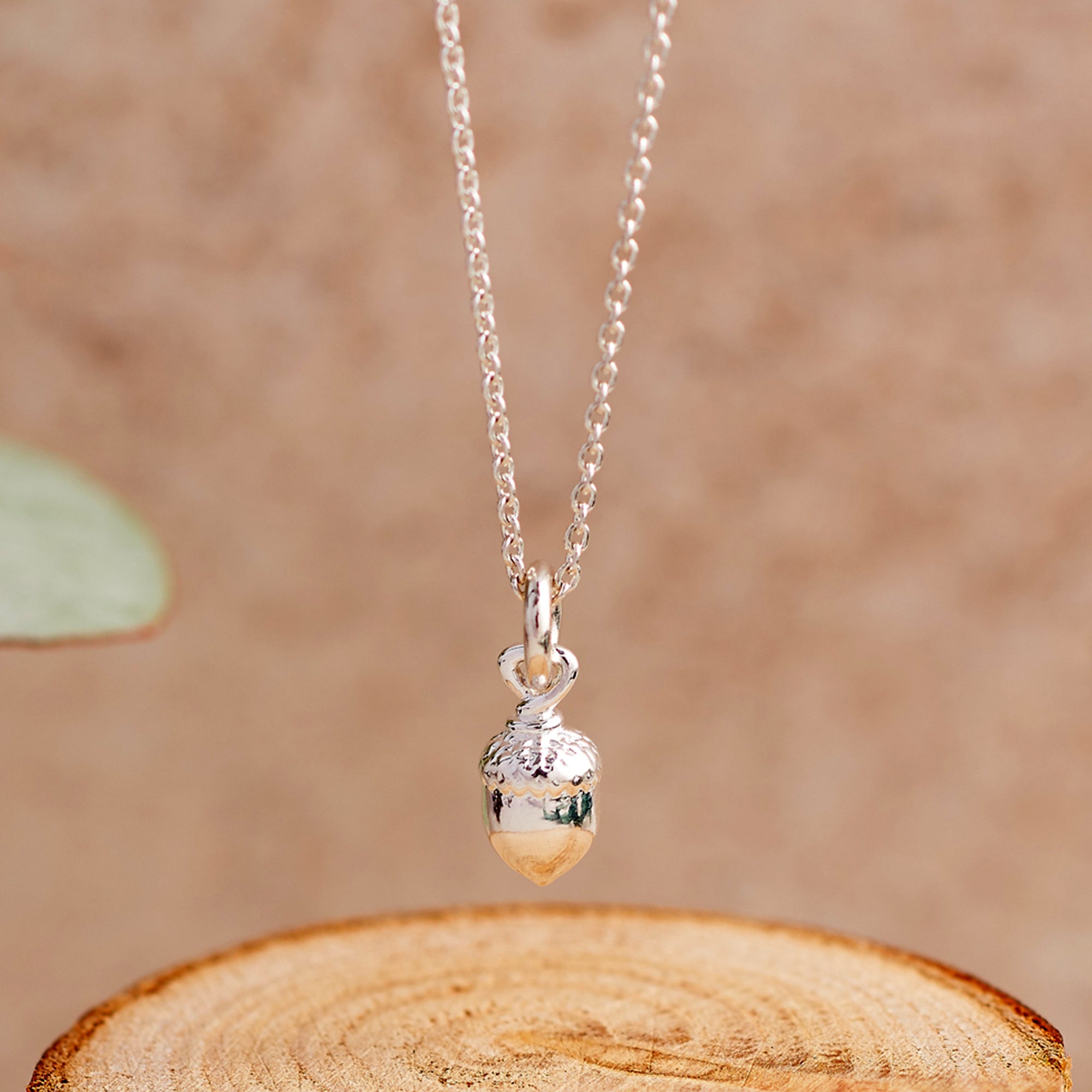 A silver acorn pendant necklace hanging from a delicate silver chain, displayed on a rustic wooden slice prop against a warm, textured beige background.