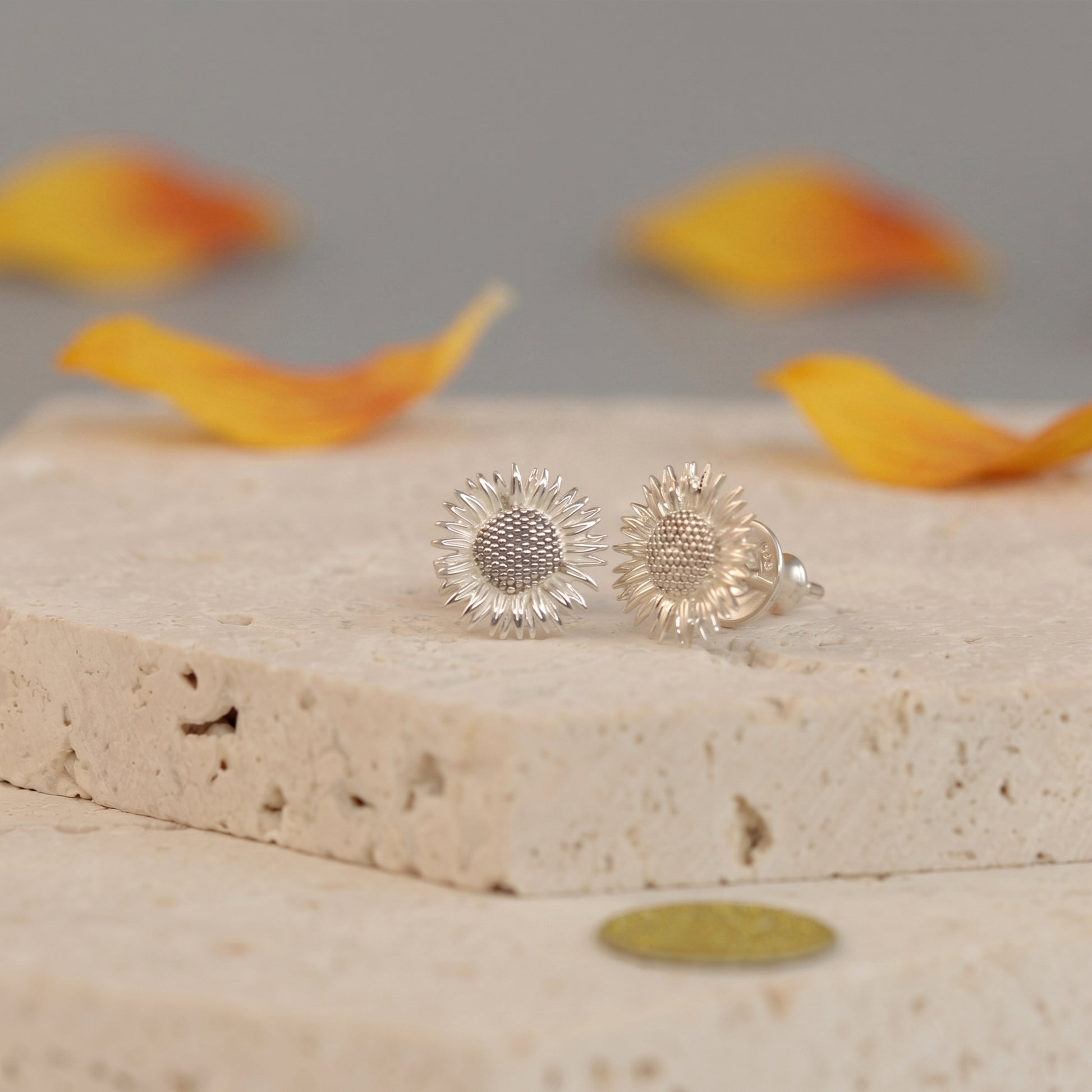 Silver sunflower-shaped earrings on a stone surface with blurred orange petals in the background