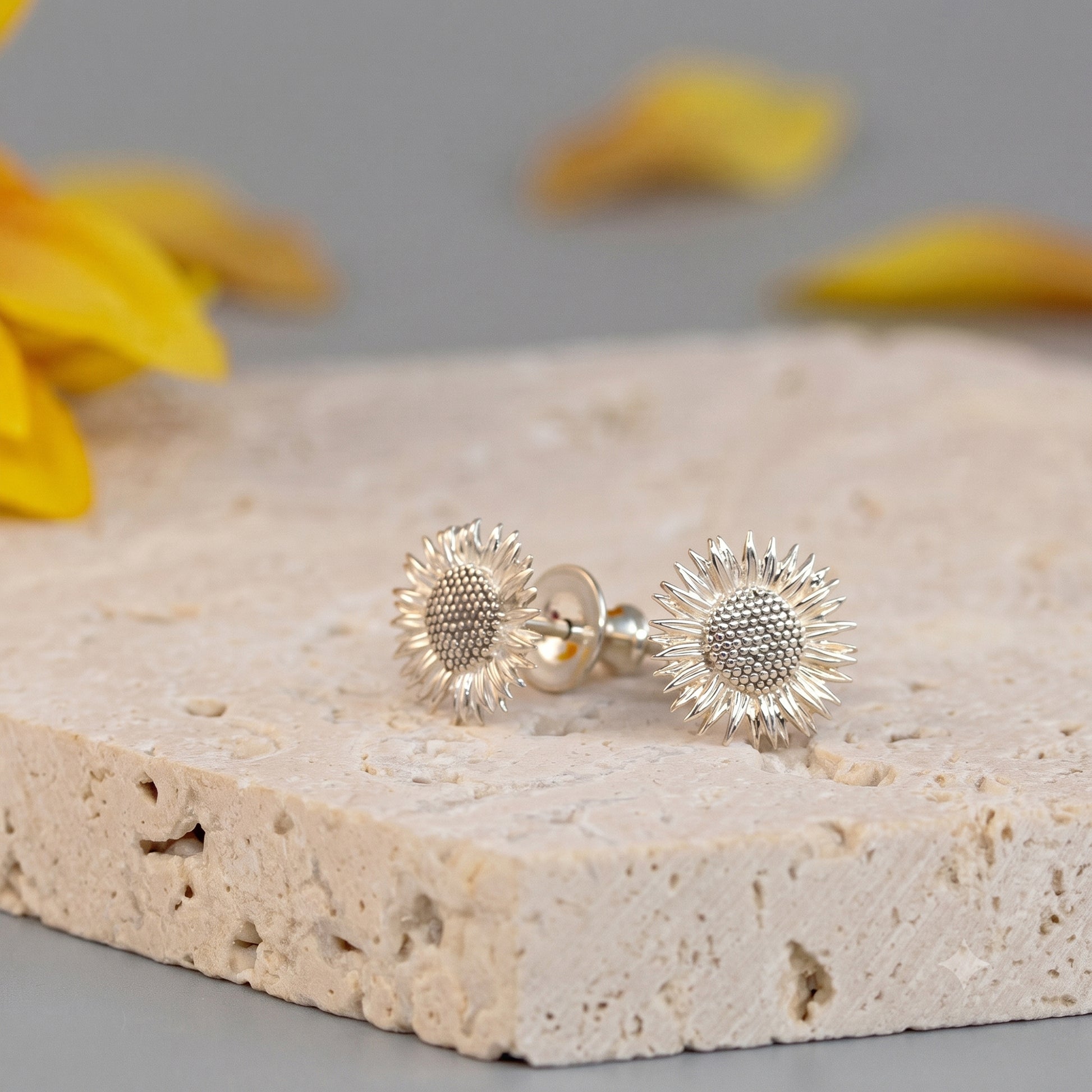 Silver sunflower earrings on a textured stone surface with blurred yellow flowers in the background.