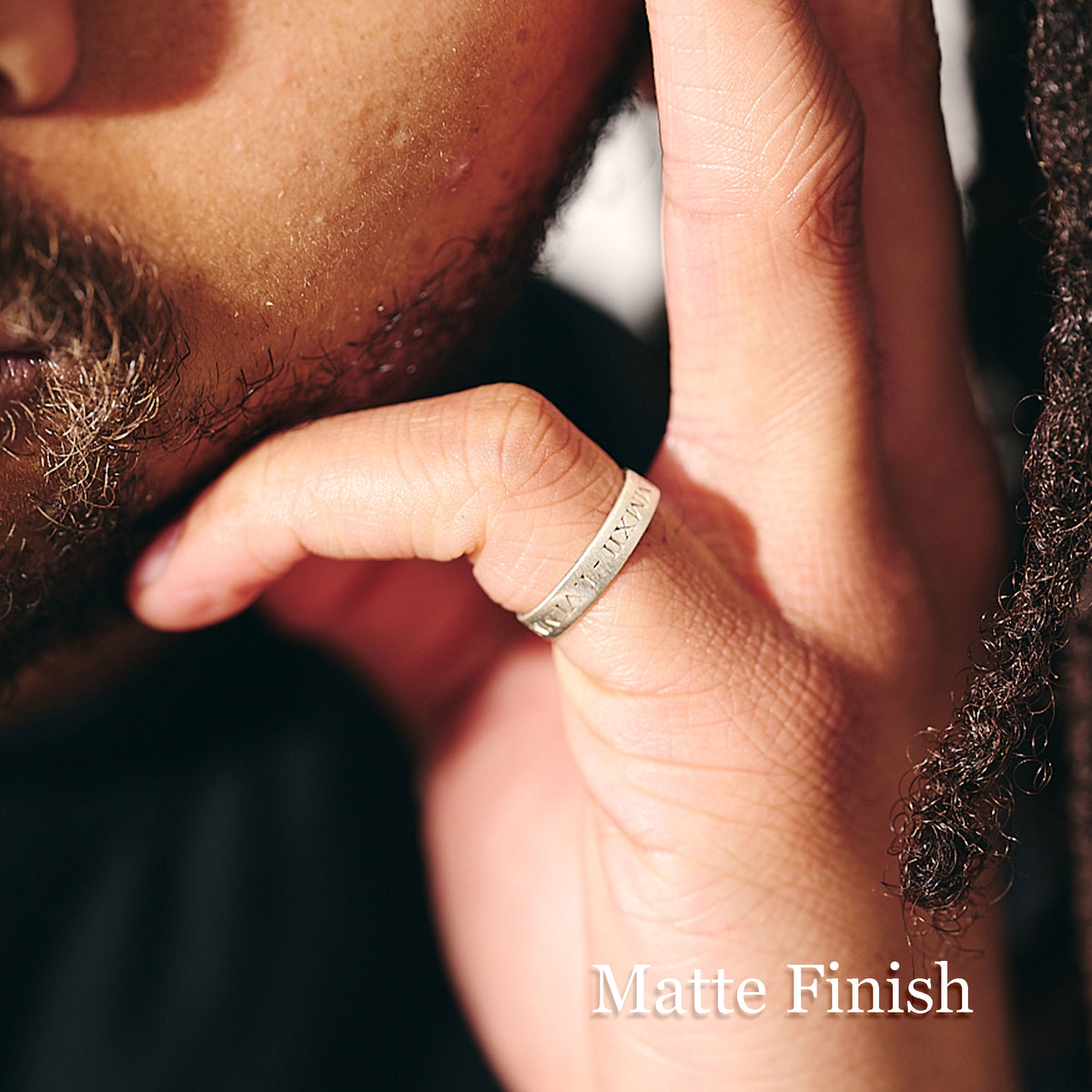 A close-up lifestyle shot of a person's hand wearing a matte silver band ring engraved with roman numerals. The image features warm, direct sunlight casting shadows, and the text 'Matte Finish' is displayed in the bottom right corner.