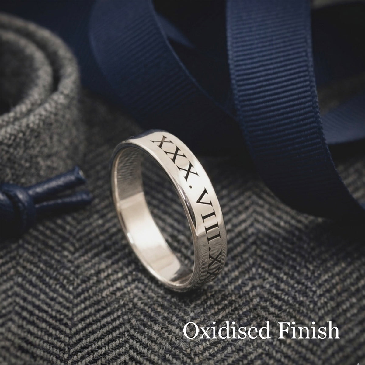 A silver band ring engraved with Roman numerals, featuring an oxidised finish, resting on a textured gray herringbone fabric with navy blue ribbons.