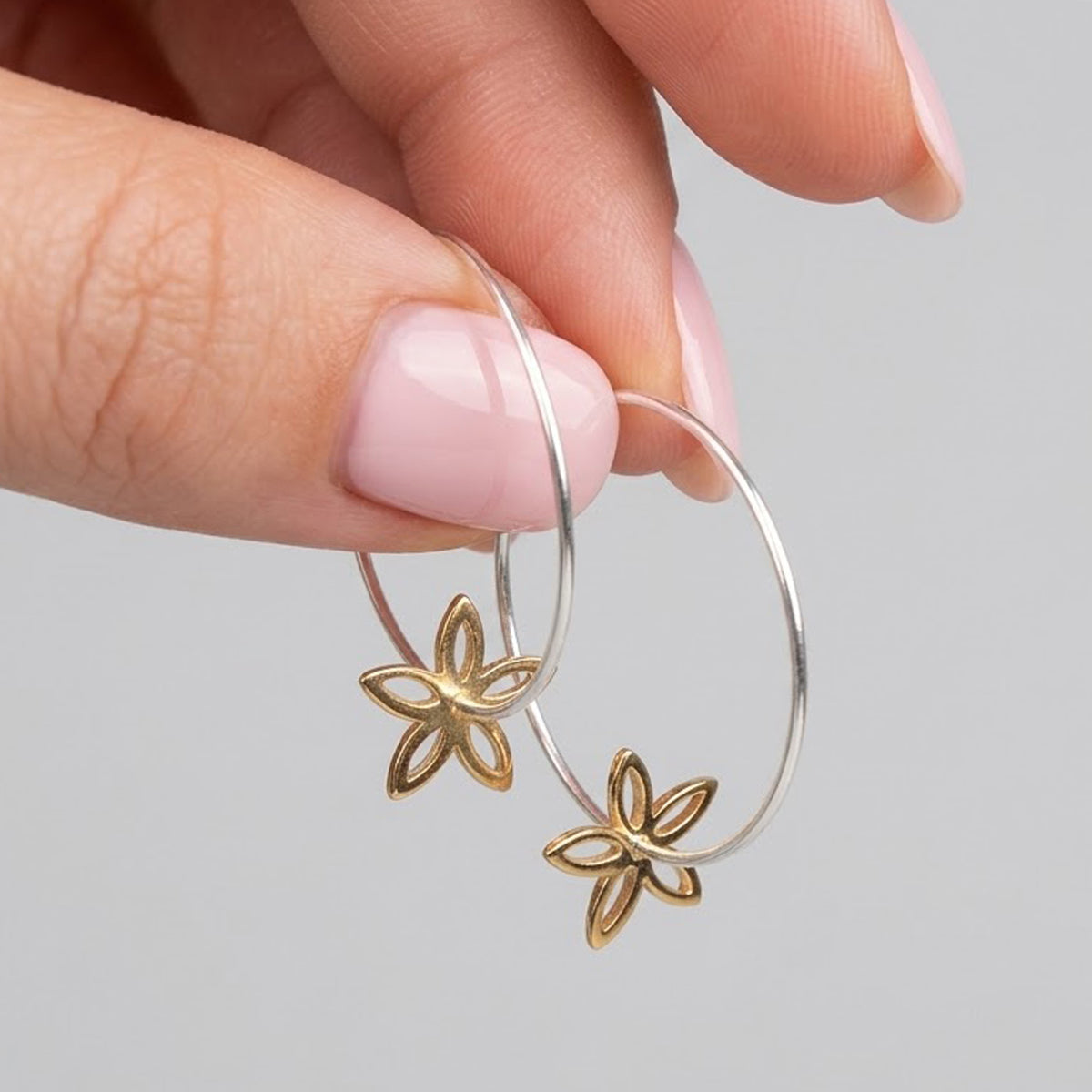 A pair of silver-tone hoop earrings with a small gold-tone flower charm hanging from the bottom of each hoop, shown in someones fingertips