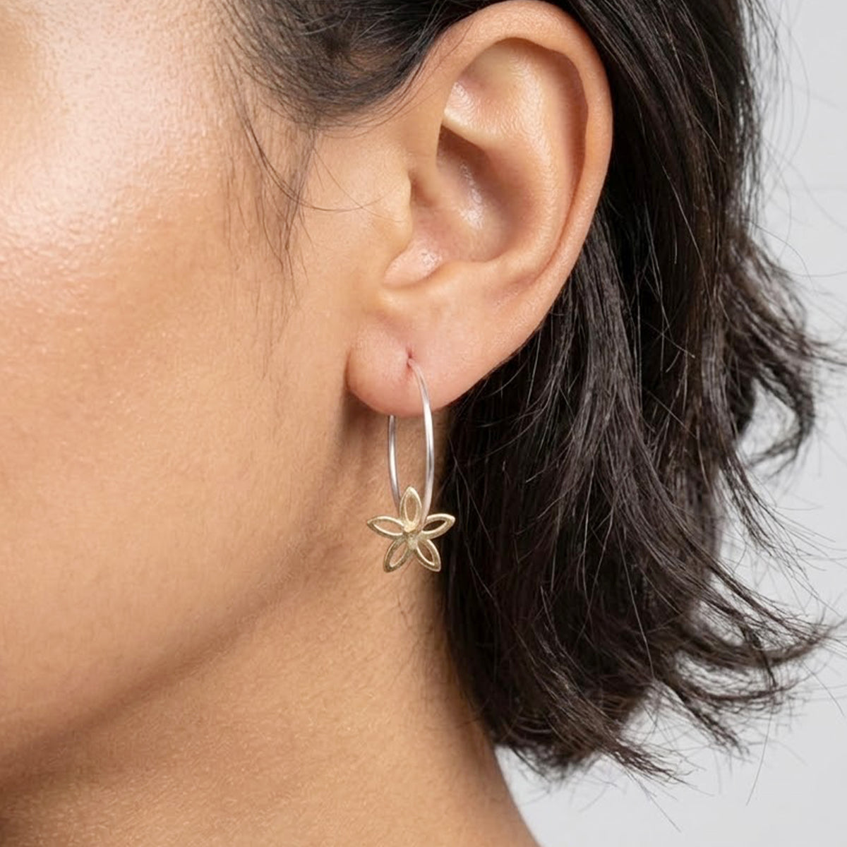 A pair of silver-tone hoop earrings with a small gold-tone flower charm hanging from the bottom of each hoop, shown in someones ears