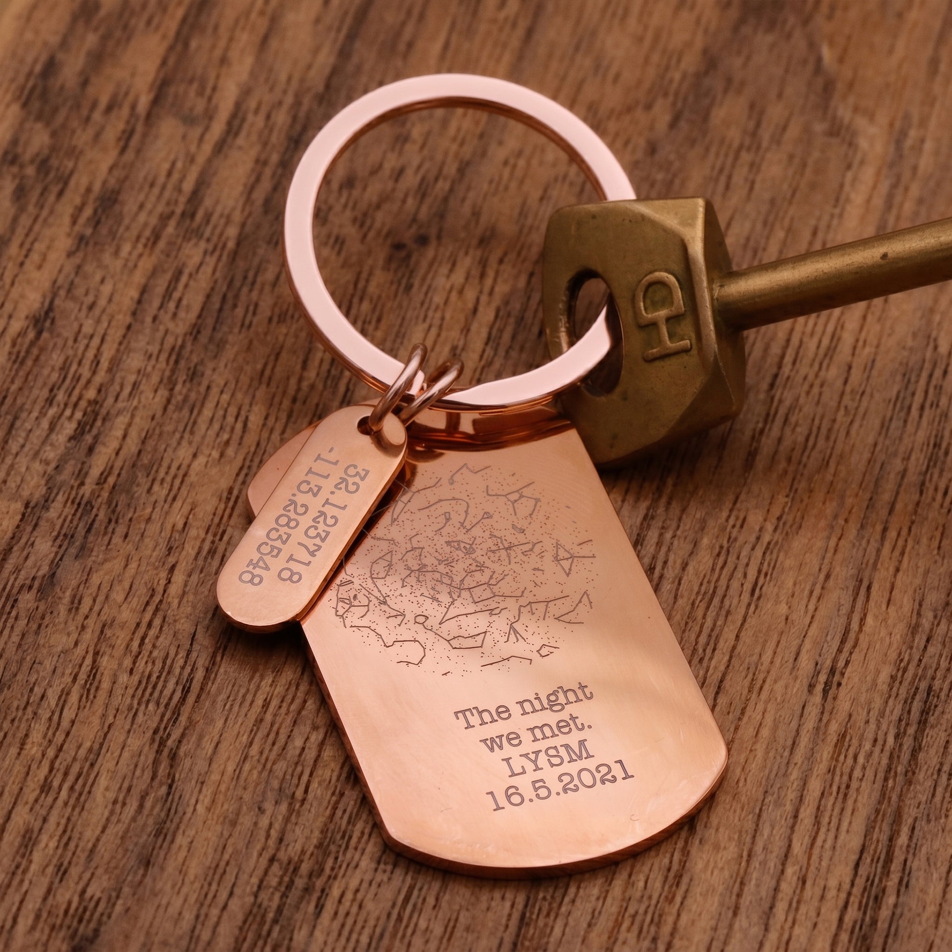 A close-up of a copper-colored engraved keyring with two tags and a brass key resting on a wooden surface. One tag is engraved with a map outline and the other with the text 'The night we met.