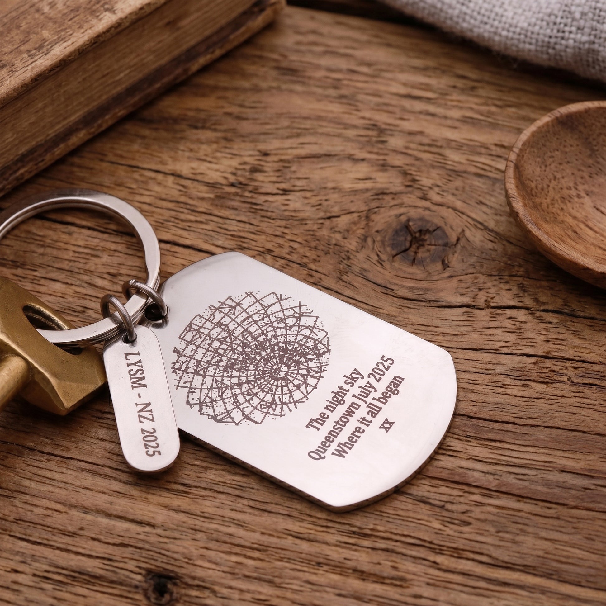 A silver metal keychain with a rounded rectangular tag and a small hanging tag, engraved with a compass design and text, displayed on a rustic wooden surface next to a brass key.