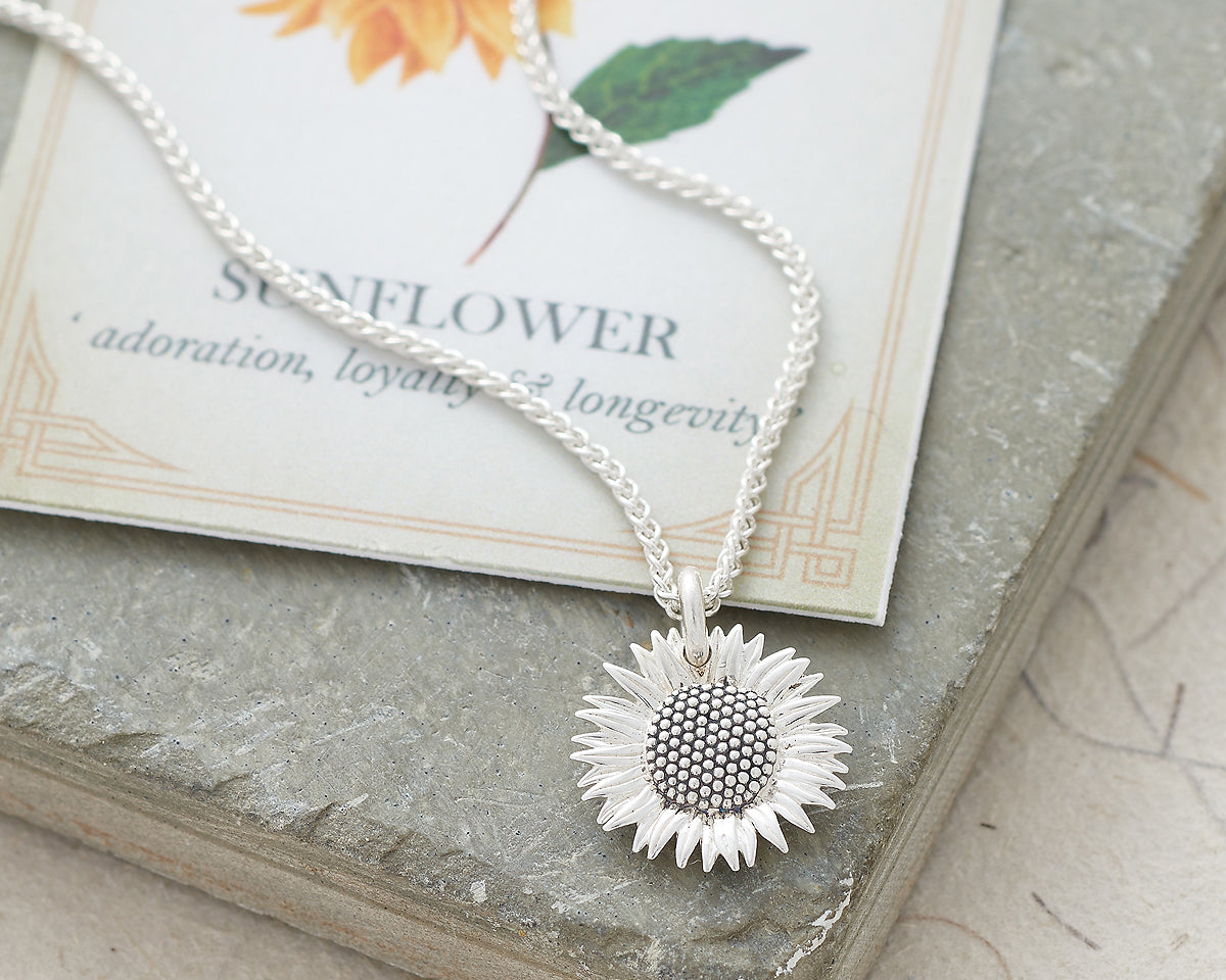 A silver sunflower pendant necklace on a delicate silver chain, resting on a textured gray stone slab next to a decorative card that reads 'SUNFLOWER adoration, loyalty & longevity'.