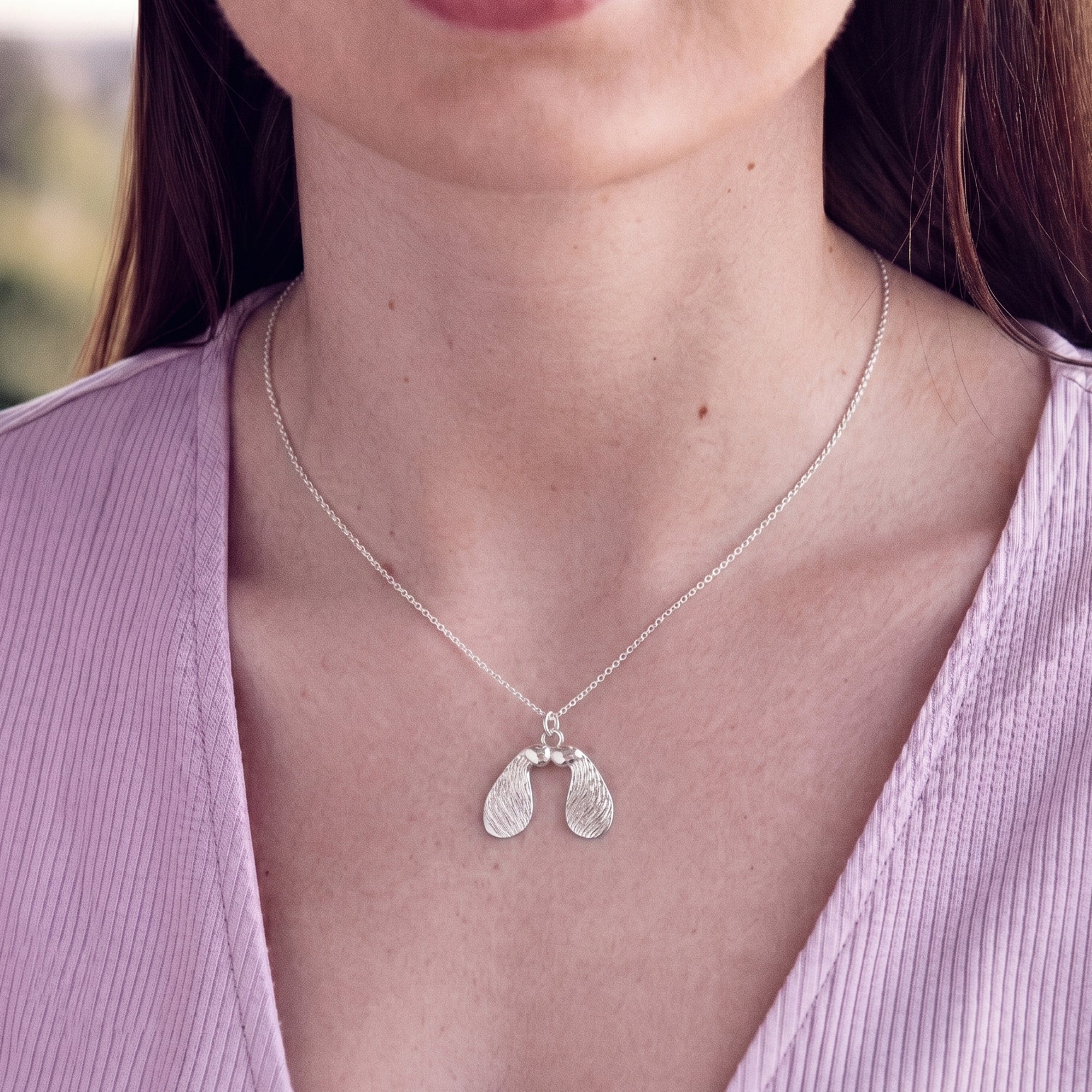 Silver necklace with a sycamore seed pendant worn by a person in a light purple top.