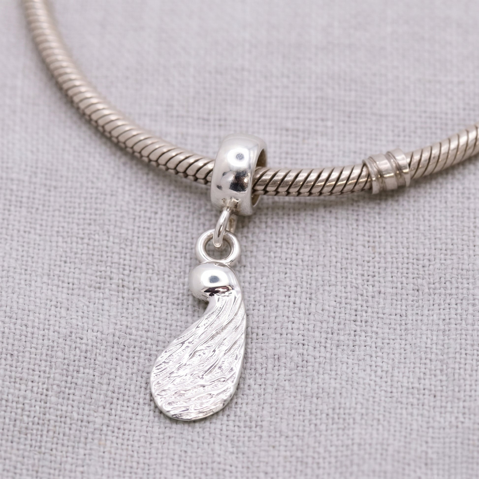 Silver charm with a seed design on a silver snake chain bracelet against a gray fabric background.