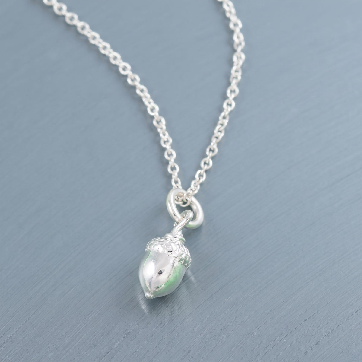 A silver chain necklace with a small acorn pendant, photographed on a textured blue-gray background.