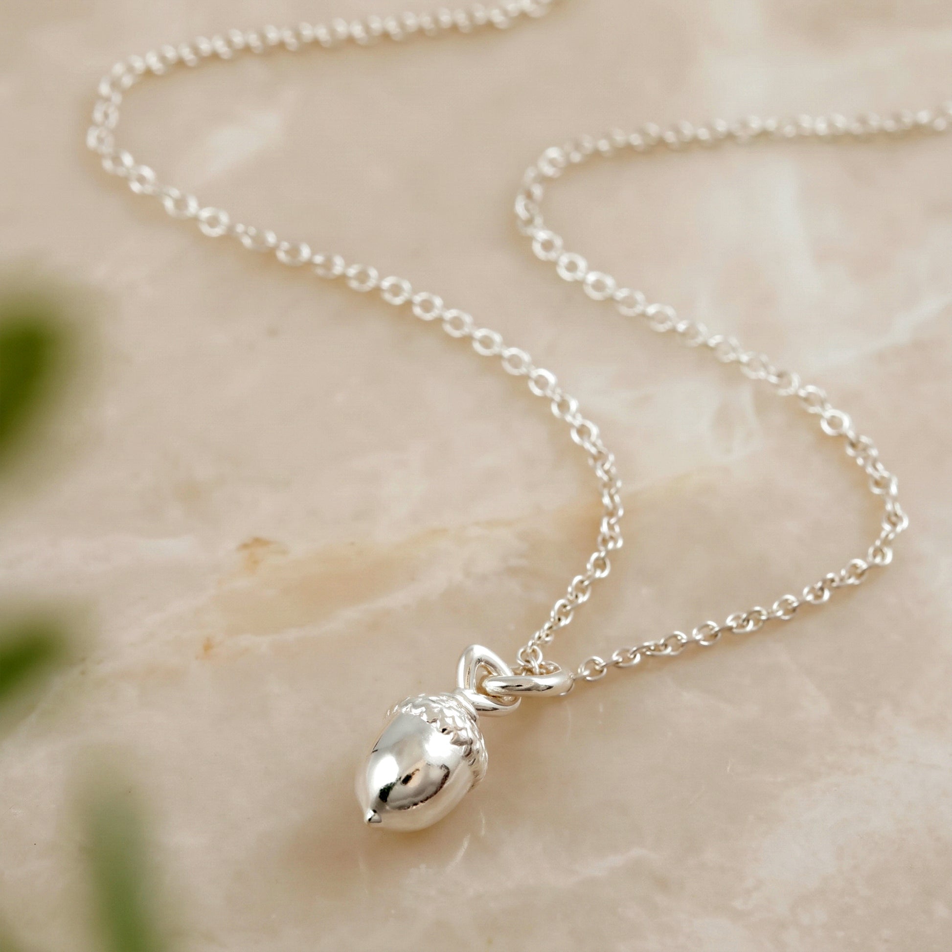 A silver acorn pendant necklace on a delicate silver chain, displayed on a light beige marble surface.