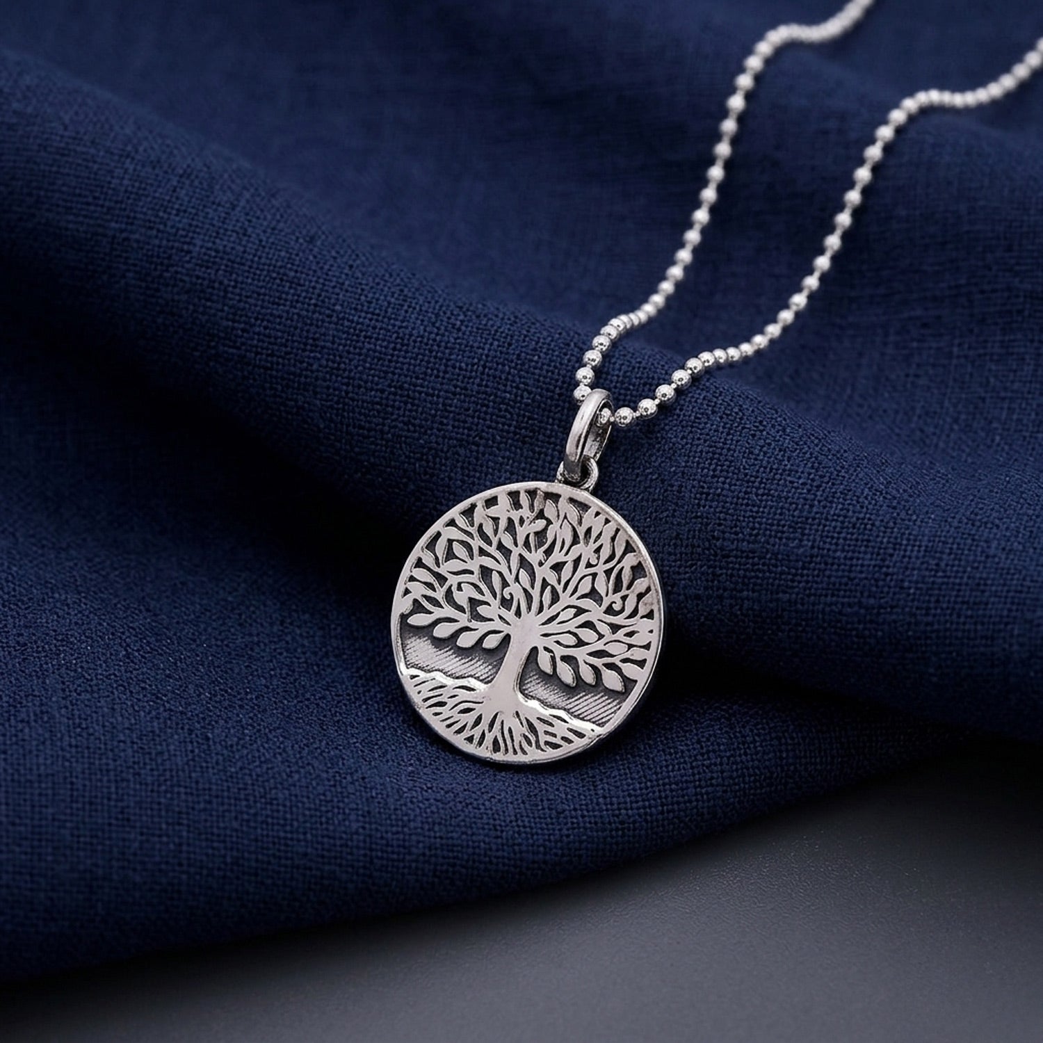 A silver tree of life pendant necklace on a matching ball chain, displayed on a textured navy blue fabric background.